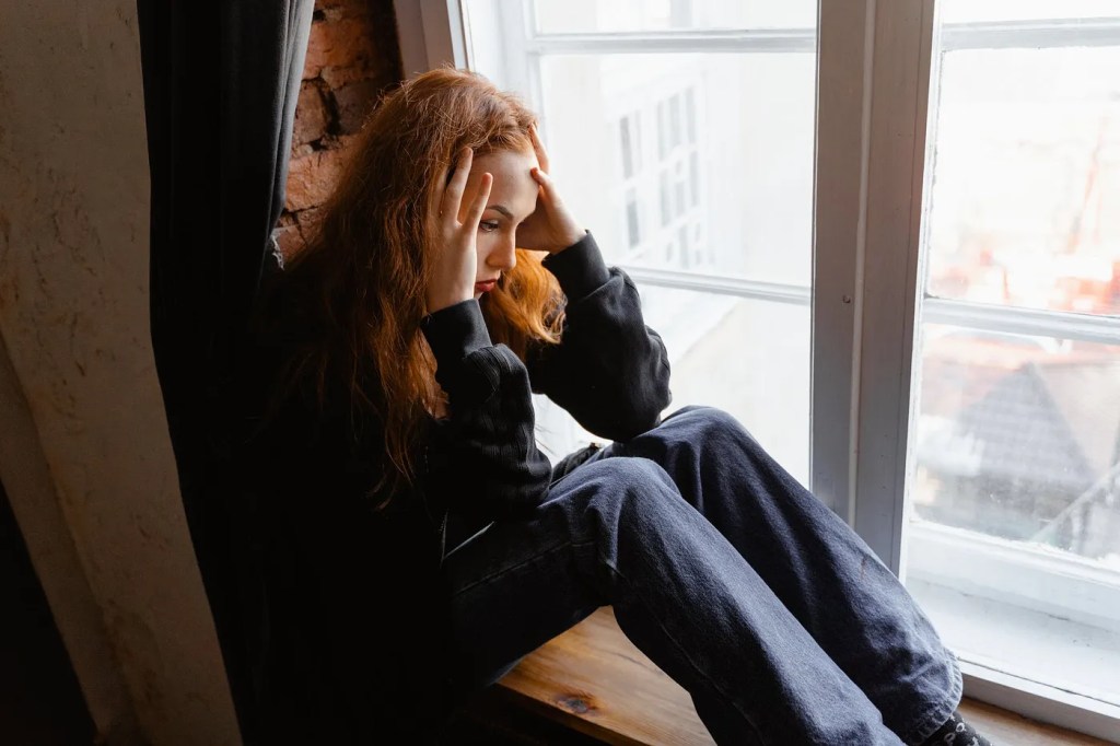 Stressed woman sitting on window sill. Photo courtesy: Mart Production / Pexel.